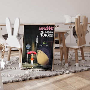 May include: A framed poster of the animated film "My Neighbor Totoro" is displayed on a rug. The poster features a large Totoro character, a girl with a red umbrella, and Japanese text. The poster is propped up against a white chair.