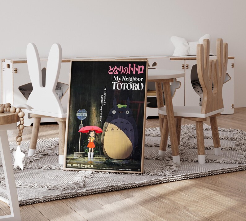 May include: A framed poster of the animated film "My Neighbor Totoro" is displayed on a rug. The poster features a large Totoro character, a girl with a red umbrella, and Japanese text. The poster is propped up against a white chair.