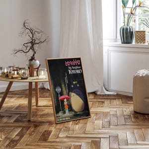 May include: A framed poster of the animated film "My Neighbor Totoro" in a room setting. The poster features a girl with a red umbrella and the character Totoro. The poster is in a wooden frame and leans against a wall. The room has a wooden floor and a round table.