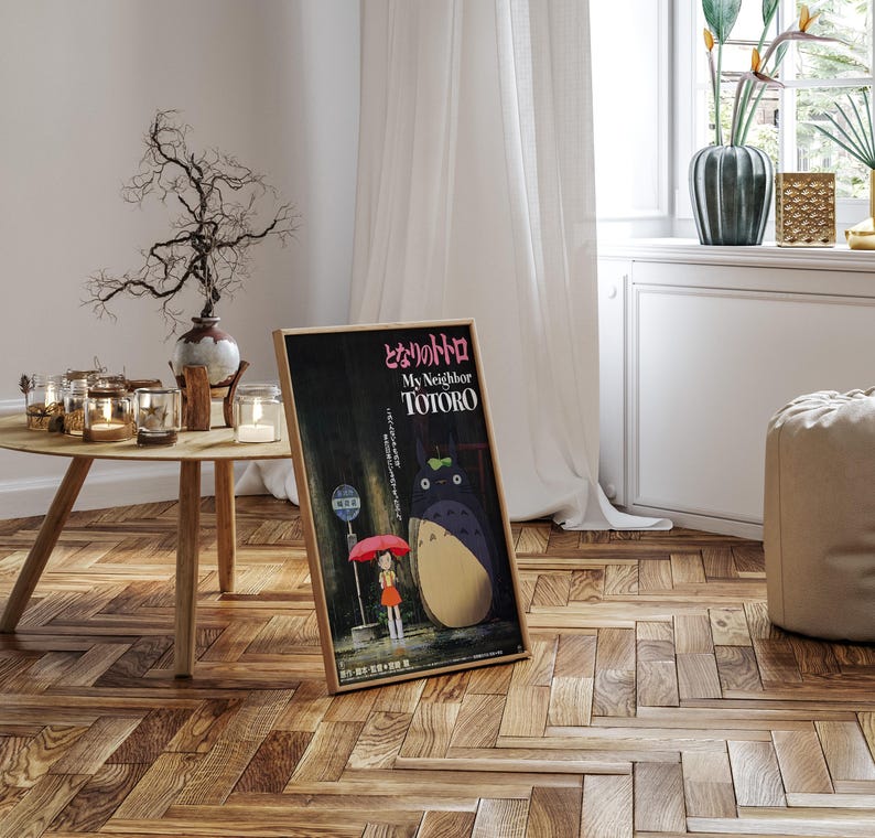May include: A framed poster of the animated film "My Neighbor Totoro" in a room setting. The poster features a girl with a red umbrella and the character Totoro. The poster is in a wooden frame and leans against a wall. The room has a wooden floor and a round table.
