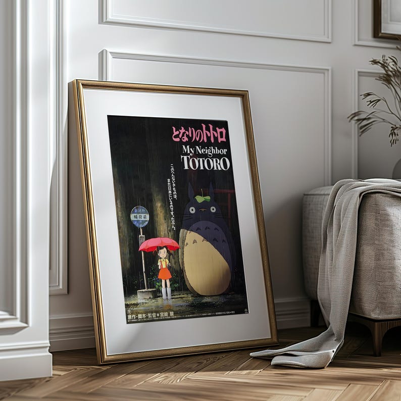 May include: Framed poster of the animated film "My Neighbor Totoro." The artwork features Totoro, a young girl with a red umbrella, and a bus stop in a rainy scene. The poster has Japanese text and is in a gold frame.