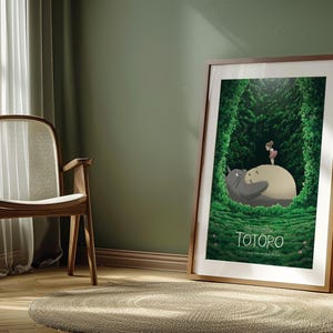 May include: Framed poster featuring a scene from "My Neighbor Totoro." The artwork depicts Totoro and a small girl in a lush green forest setting. The poster is in a wooden frame and leans against a green wall.
