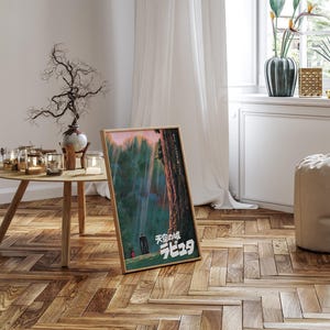 May include: A framed poster with Japanese text and a nature scene featuring a waterfall and two figures. The poster is leaning against a wall on a wooden floor. The room has a neutral color palette with a round wooden table and a vase.