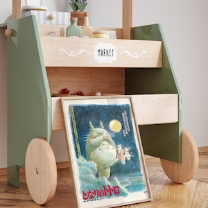 Puede incluir: Un carrito de mercado de juguete de madera con detalles en verde y madera natural. El carrito presenta un p&oacute;ster enmarcado de un personaje de dibujos animados, una estanter&iacute;a con objetos decorativos y la palabra "MARKET" en blanco.
