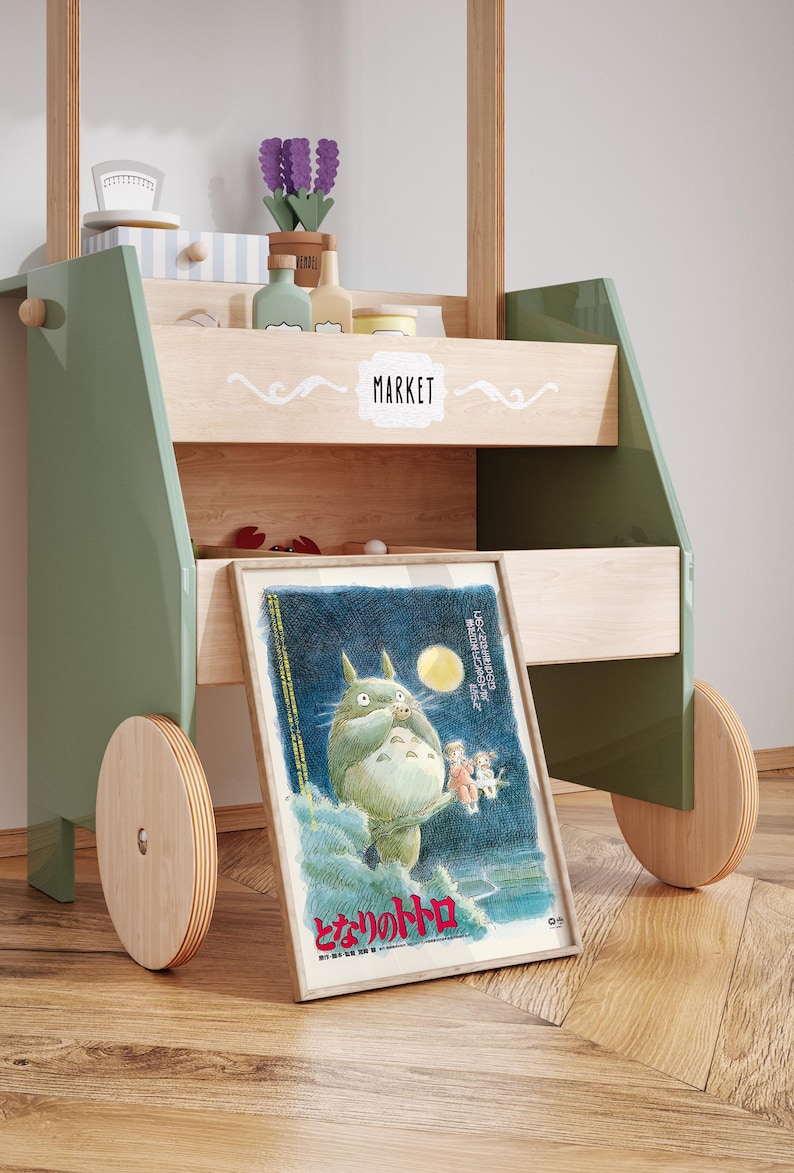 Puede incluir: Un carrito de mercado de juguete de madera con detalles en verde y madera natural. El carrito presenta un p&oacute;ster enmarcado de un personaje de dibujos animados, una estanter&iacute;a con objetos decorativos y la palabra "MARKET" en blanco.