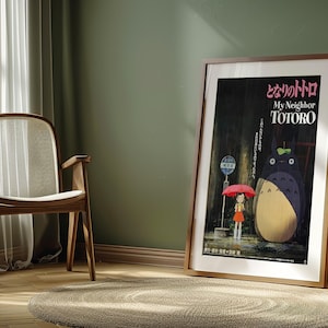 May include: Framed poster of the animated film "My Neighbor Totoro" in a room setting. The poster features the film's title in Japanese and English, with an illustration of Totoro and a girl under an umbrella. The room has a wooden chair, a round rug, and a green wall.