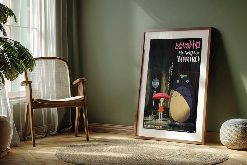 May include: Framed poster of the animated film "My Neighbor Totoro" in a room setting. The poster features the film's title in Japanese and English, with an illustration of Totoro and a girl under an umbrella. The room has a wooden chair, a round rug, and a green wall.