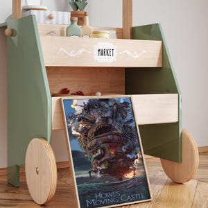 Op de afbeelding: Een houten speelgoedmarktstand met een groen frame en houten wielen. De stand heeft planken met miniatuur speelgoed en een ingelijste poster van Howl's Moving Castle. Het woord "MARKET" staat op de voorkant.