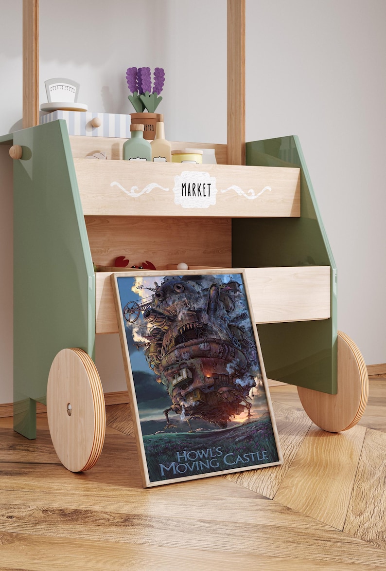 Op de afbeelding: Een houten speelgoedmarktstand met een groen frame en houten wielen. De stand heeft planken met miniatuur speelgoed en een ingelijste poster van Howl's Moving Castle. Het woord "MARKET" staat op de voorkant.