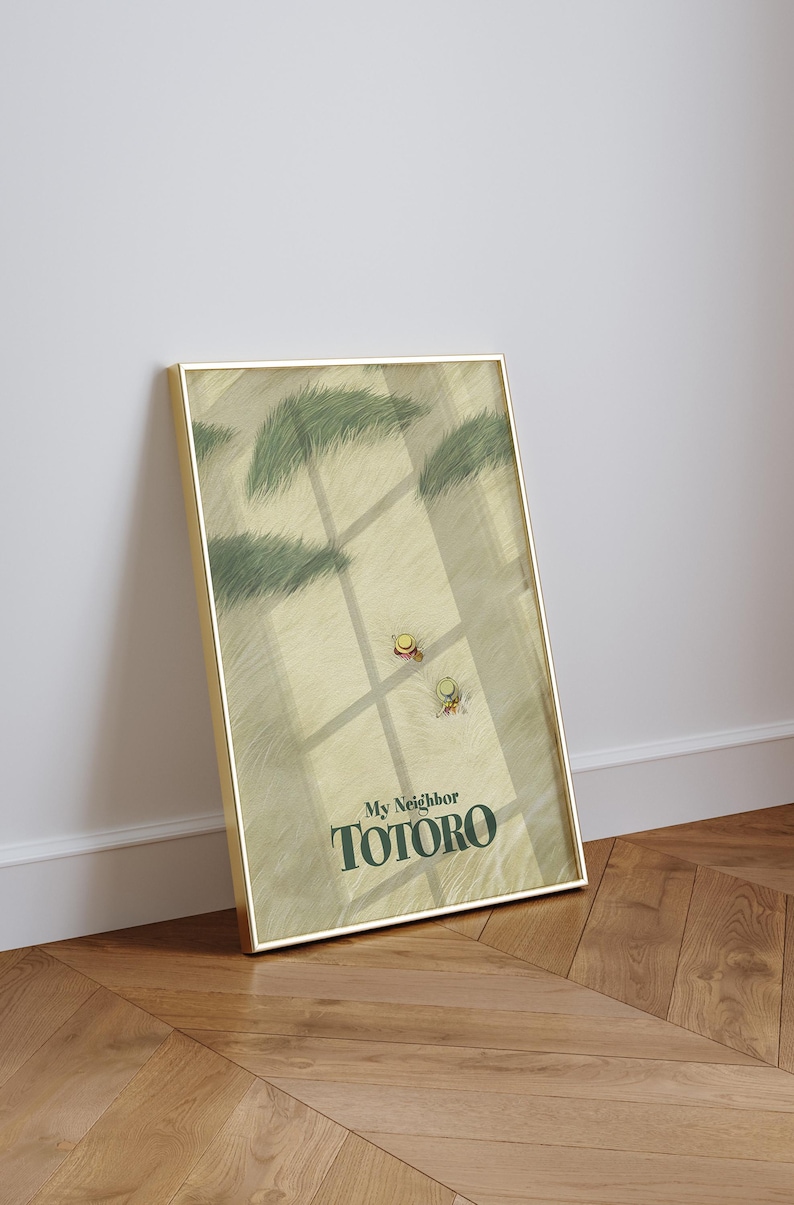 May include: A framed art print featuring a scene from "My Neighbor Totoro." The artwork depicts a field with two small figures wearing hats, with the title "My Neighbor Totoro" in green text at the bottom. The frame is gold.