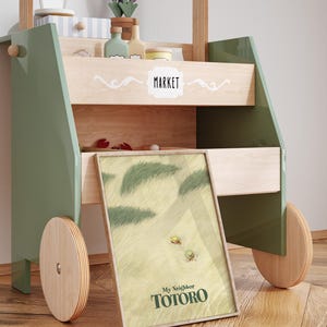 May include: A wooden toy market cart with green and natural wood accents. The cart features a framed "My Neighbor Totoro" print, shelves with toy accessories, and wooden wheels. The word "MARKET" is displayed on the cart.