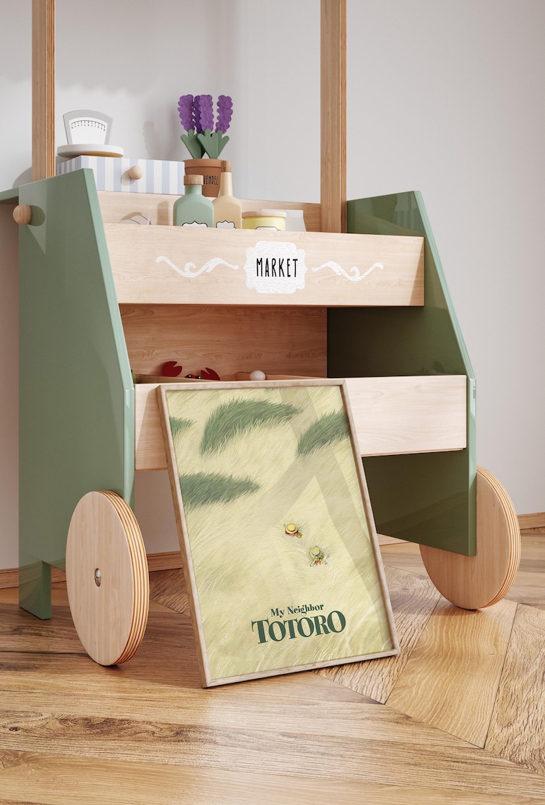 May include: A wooden toy market cart with green and natural wood accents. The cart features a framed "My Neighbor Totoro" print, shelves with toy accessories, and wooden wheels. The word "MARKET" is displayed on the cart.