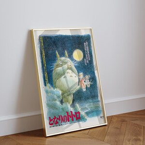 Puede incluir: P&oacute;ster enmarcado de la pel&iacute;cula de animaci&oacute;n "Mi vecino Totoro". La obra de arte representa a Totoro, un gran esp&iacute;ritu del bosque, con dos ni&ntilde;os y un cielo nocturno iluminado por la luna. El p&oacute;ster est&aacute; en un marco dorado y se apoya en una pared blanca.