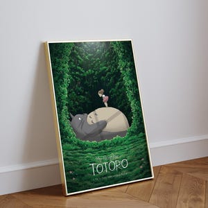 May include: Framed art print featuring a scene from "My Neighbor Totoro." The artwork depicts Totoro lying down with a girl standing on his belly, surrounded by lush green foliage. The title "TOTORO" is displayed at the bottom.