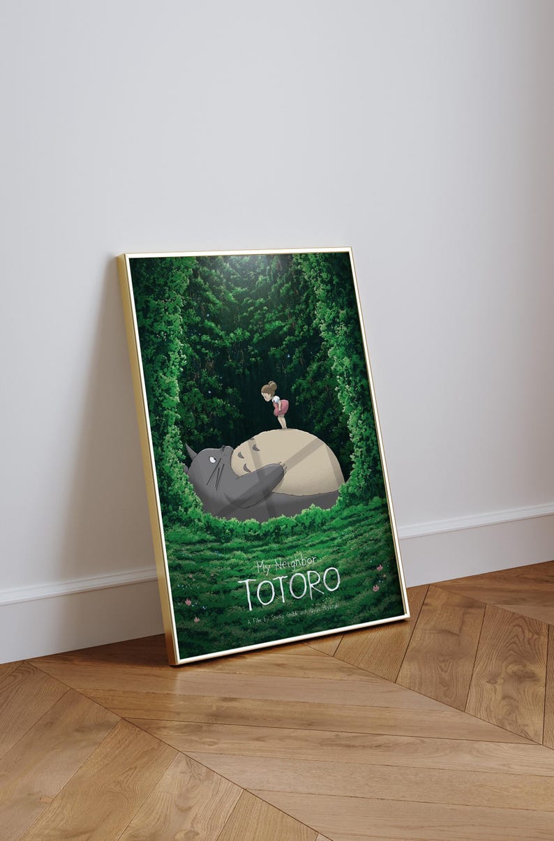 May include: Framed art print featuring a scene from "My Neighbor Totoro." The artwork depicts Totoro lying down with a girl standing on his belly, surrounded by lush green foliage. The title "TOTORO" is displayed at the bottom.
