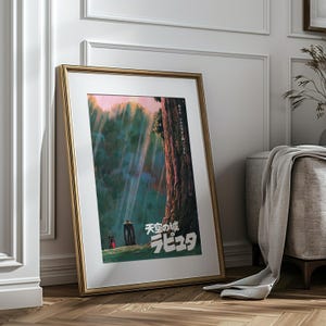 May include: Framed poster featuring a scene from the animated film "Castle in the Sky." The artwork depicts a forest with a large tree, a figure, and the Japanese title. The frame is gold-colored, and the poster is leaning against a white wall and wooden floor.