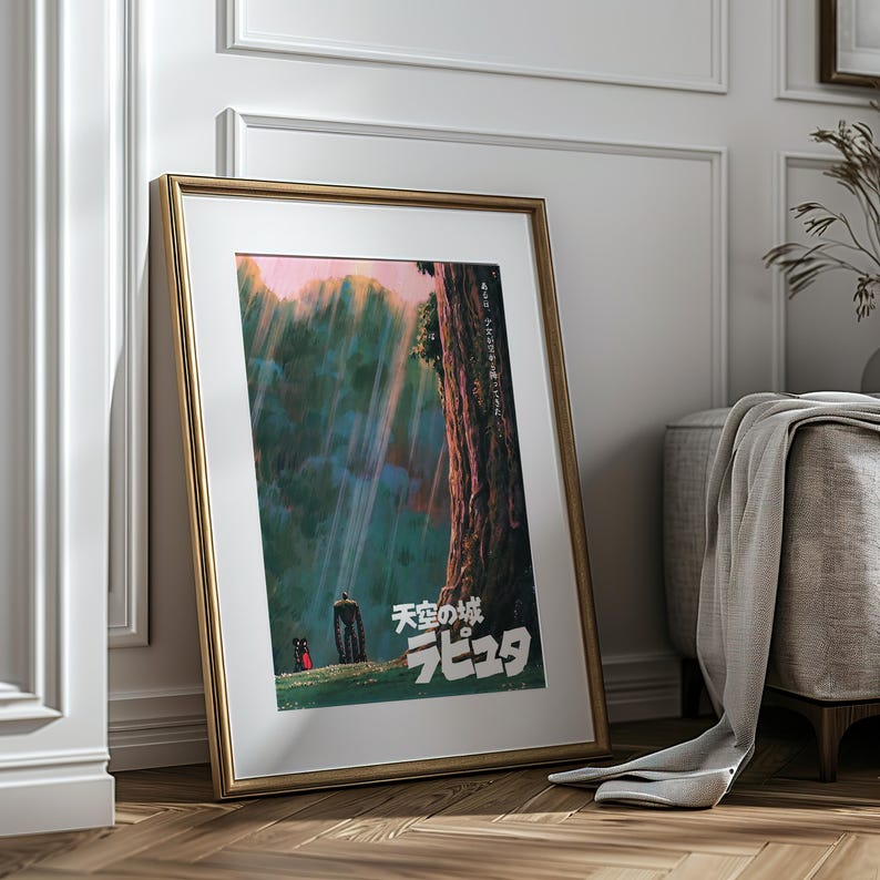 May include: Framed poster featuring a scene from the animated film "Castle in the Sky." The artwork depicts a forest with a large tree, a figure, and the Japanese title. The frame is gold-colored, and the poster is leaning against a white wall and wooden floor.