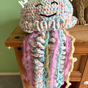 May include: A crochet jellyfish toy with the text Jumbo Jellyfish. The head is made of white yarn with multicolored speckles and a black stitched smiling face. It features long, dangling tentacles in shades of pink, yellow, and light blue.