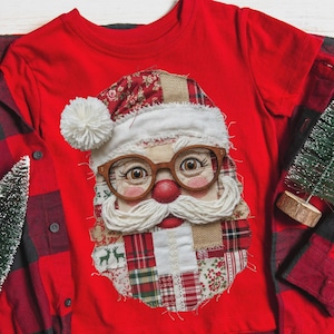 Può includere: Una t-shirt rosso acceso con una grande applicazione patchwork centrale raffigurante il volto di Babbo Natale. Il design include un pon-pon di lana bianca, baffi di lana bianca, occhiali in legno e vari tessuti con motivi rossi e verdi.