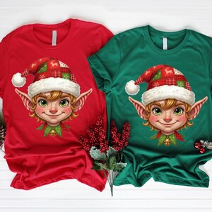 Op de afbeelding: Twee T-shirts met korte mouwen, één rode en één groene, elk met een cartoon elfengezicht. De elf draagt een rood-groene geruite hoed met een witte pompon en een groene kraag met een bel. De elf heeft blond haar en groene ogen.