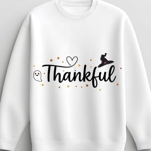 May include: White sweatshirt featuring the word "Thankful" in black script, with a ghost, heart, and witch hat design. Gold dots surround the text. A seasonal design for autumn.
