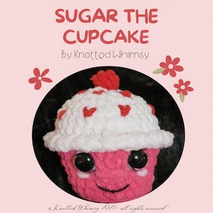Könnte beinhalten: Ein handgehäkelter rosa und weißer Cupcake mit großen schwarzen Augen und einem lächelnden Gesicht. Der Cupcake ist mit einem roten Pompon und roten Herzakzenten verziert. Der Text "SUGAR THE CUPCAKE" steht über dem Cupcake.
