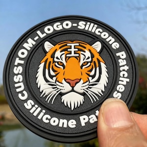 Op de afbeelding: Een ronde zwarte siliconen patch met een centraal oranje, wit en zwart tijgerkopmotief. De buitenrand bevat de verhoogde witte tekst "CUSTOM-LOGO-Silicone Patches" die herhaald wordt rond de omtrek van het embleem.