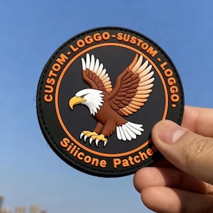 Op de afbeelding: Een ronde zwarte siliconen patch met een reliëf van een adelaar met bruine vleugels, een witte kop en een gele snavel. De rand bevat de oranje tekst CUSTOM-LOGGO-SUSTOM-LOGGO en Silicone Patches in een vetgedrukt, schreefloos lettertype.