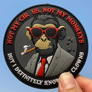 Op de afbeelding: Een ronde zwarte patch met een cartoon chimpansee in een grijs pak, rode das en een zonnebril met rode randen, terwijl hij een sigaar rookt. De tekst luidt NOT MY CIRCUS, NOT MY MONKEYS, BUT I DEFINITELY KNOW THE CLOWNS, met het getal 51.