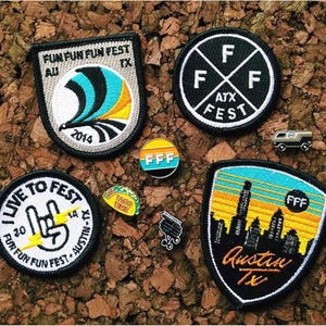 May include: A collection of embroidered patches and pins with the theme of a music festival. The patches feature text like "FUN FUN FUN FEST" and "I LIVE TO FEST". The pins include a taco, a van, and a pin with the letters "FFF".