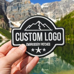 May include: A black embroidered patch with a white border and white stitching. The design features a mountain outline above the text CUSTOM LOGO and EMBROIDERY PATCHES, with three stars centered at the bottom. The patch has a rounded rectangular shape.