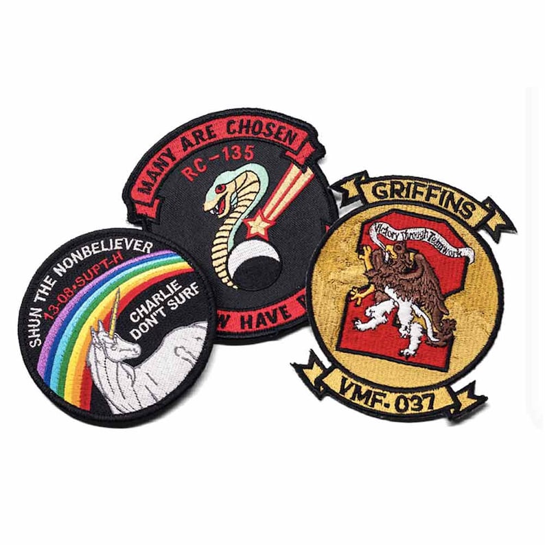 May include: Three embroidered patches with various designs. One patch features a cobra and the text "MANY ARE CHOSEN RC-135". Another shows a unicorn with a rainbow and the text "SHUN THE NONBELIEVER". The third patch displays a griffon and the text "GRIFFINS VMF-037".