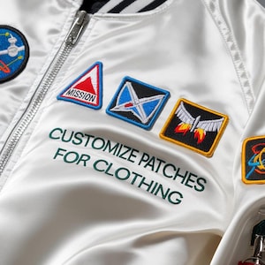 May include: White satin bomber jacket with embroidered patches. Patches include a red and white triangle with "MISSION" text, a blue and white design, and a rocket ship. The text "CUSTOMIZE PATCHES FOR CLOTHING" is embroidered in green.