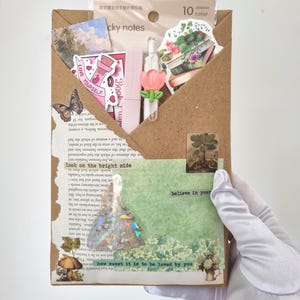 Puede incluir: Un sobre de papel marrón lleno de pegatinas, notas adhesivas y elementos decorativos. El sobre contiene una variedad de pegatinas, incluyendo diseños florales y de mariposas, y el texto "believe in your" y "look on the bright side".