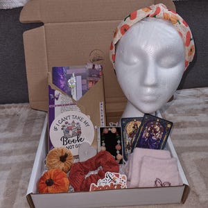 May include: A gift box filled with various items, including a headband with pumpkin print, a book-themed coaster, a scrunchie, a bracelet, two tarot cards, a pair of pink socks, and two small orange pumpkins. The box is open, revealing the contents.