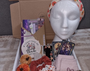 May include: A gift box filled with various items, including a headband with pumpkin print, a book-themed coaster, a scrunchie, a bracelet, two tarot cards, a pair of pink socks, and two small orange pumpkins. The box is open, revealing the contents.