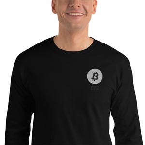 Könnte beinhalten: Schwarzes Langarmshirt mit einem Bitcoin-Logo auf der linken Brust. Das Logo ist ein silbernes Bitcoin-Symbol in einem Kreis, darunter steht "BTC". Das Shirt ist aus weichem Material.