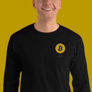 Könnte beinhalten: Schwarzes Langarmshirt mit einem gelben Bitcoin-Symbol und den Buchstaben "BTC" auf der Brust gestickt. Das Shirt besteht aus einem weichen Material und eignet sich für Freizeitkleidung.