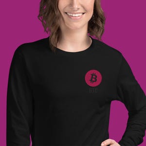 Könnte beinhalten: Schwarzes Langarmshirt mit einem roten Bitcoin-Symbol in einem Kreis auf der Brust. Die Buchstaben "BTC" sind unter dem Symbol aufgedruckt. Das Shirt besteht aus einem weichen Material.