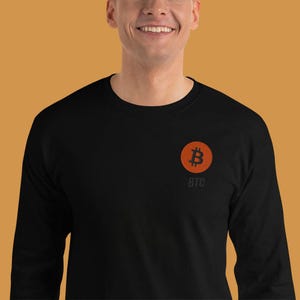 Könnte beinhalten: Schwarzes Langarmshirt mit einem orangefarbenen Bitcoin-Symbol und den Buchstaben "BTC" darunter. Das Bitcoin-Symbol ist ein orangefarbenes "B" in einem orangefarbenen Kreis. Das Shirt ist aus weichem Material.