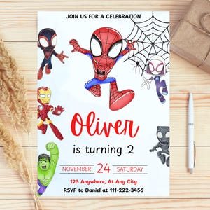 May include: A white invitation card with cartoon illustrations of Spider-Man and other superheroes. The text "Oliver is turning 2" is in red, with event details below. The card is on a wooden surface.