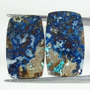 Peut inclure: Deux pierres précieuses rectangulaires aux coins arrondis. Les pierres présentent un mélange saisissant de motifs bleu foncé, marron et blanc. Les pierres sont maintenues par un outil argenté, mettant en valeur leurs surfaces polies et leurs textures naturelles.