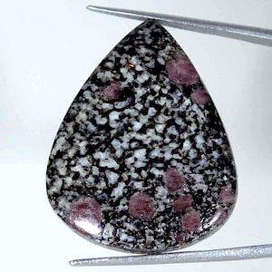 Pode incluir: Uma pedra preciosa em formato de pera com um padrão salpicado de inclusões minerais pretas e brancas por toda a superfície, acentuada por vários depósitos de cristais distintos, lisos e arredondados, de cor rosa escuro a magenta.