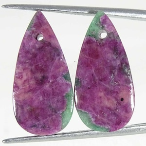 Może przedstawiać: Dwa kamienie szlachetne w kształcie łezki z otworem u góry. Kamienie mają mieszankę głębokich różowych i jasnozielonych kolorów, z wypolerowaną powierzchnią. Kamienie szlachetne są trzymane przez metalowe narzędzie, co sugeruje, że są przeznaczone do tworzenia biżuterii.