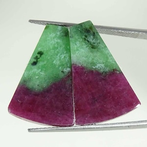 Könnte beinhalten: Zwei polierte Rubin-Zoisit-Edelsteine, gehalten von einem Metallwerkzeug. Die Steine sind dreieckig, mit einem leuchtend grünen oberen Bereich und einem tiefroten-violetten unteren Bereich. Dunkelgrüne und schwarze Einschlüsse sind sichtbar.