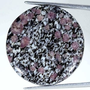 Könnte beinhalten: Ein runder Cabochon-Edelstein mit einem gesprenkelten Muster aus schwarzen und weißen Mineraleinschlüssen, durchsetzt mit zahlreichen kleinen, abgerundeten, durchscheinenden rosa Kristallen auf der polierten Oberfläche.