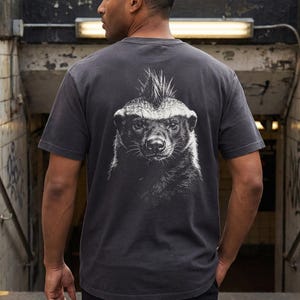 May include: Dark gray t-shirt featuring a detailed black and white illustration of a honey badger on the back. The honey badger has a mohawk-like crest. The shirt is a classic crew neck style.
