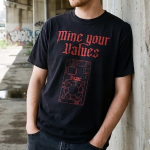 May include: A person wearing a black t-shirt with the text "Mine your Values" in red gothic lettering. The shirt features a red graphic design. The person is also wearing a tan beanie and dark blue jeans.