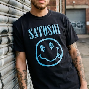 Puede incluir: Camiseta negra con la palabra "SATOSHI" en azul claro, sobre una carita sonriente con símbolos de dólar en los ojos y la lengua fuera. La persona está apoyada en una pared metálica.