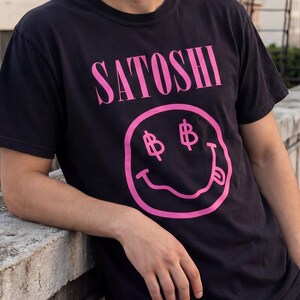 Könnte beinhalten: Schwarzes T-Shirt mit dem Wort "SATOSHI" in rosa Blockbuchstaben über einem rosa Smiley-Gesicht mit Dollarzeichen-Augen. Die Person trägt das T-Shirt und lehnt sich an eine Betonbrüstung.